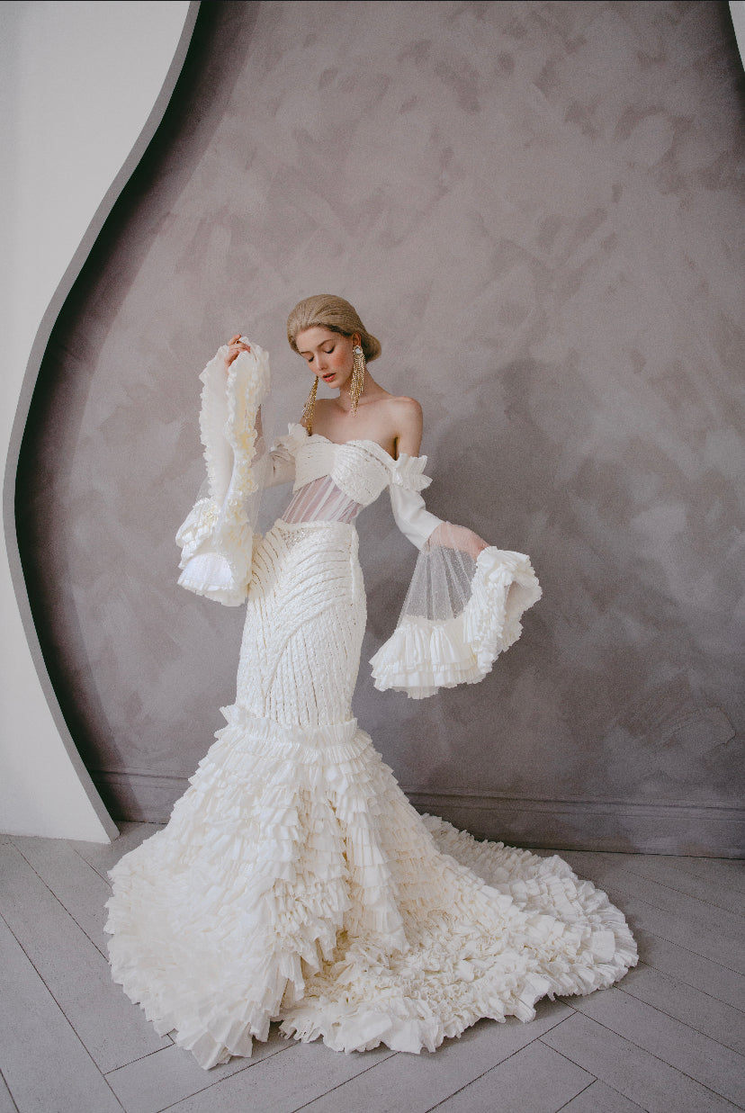 A model wears Zoba Martin's custom toilet tissue dress for breast cancer awareness: an off-shoulder mermaid gown with pleated ruffle tiers and sheer bell sleeves.